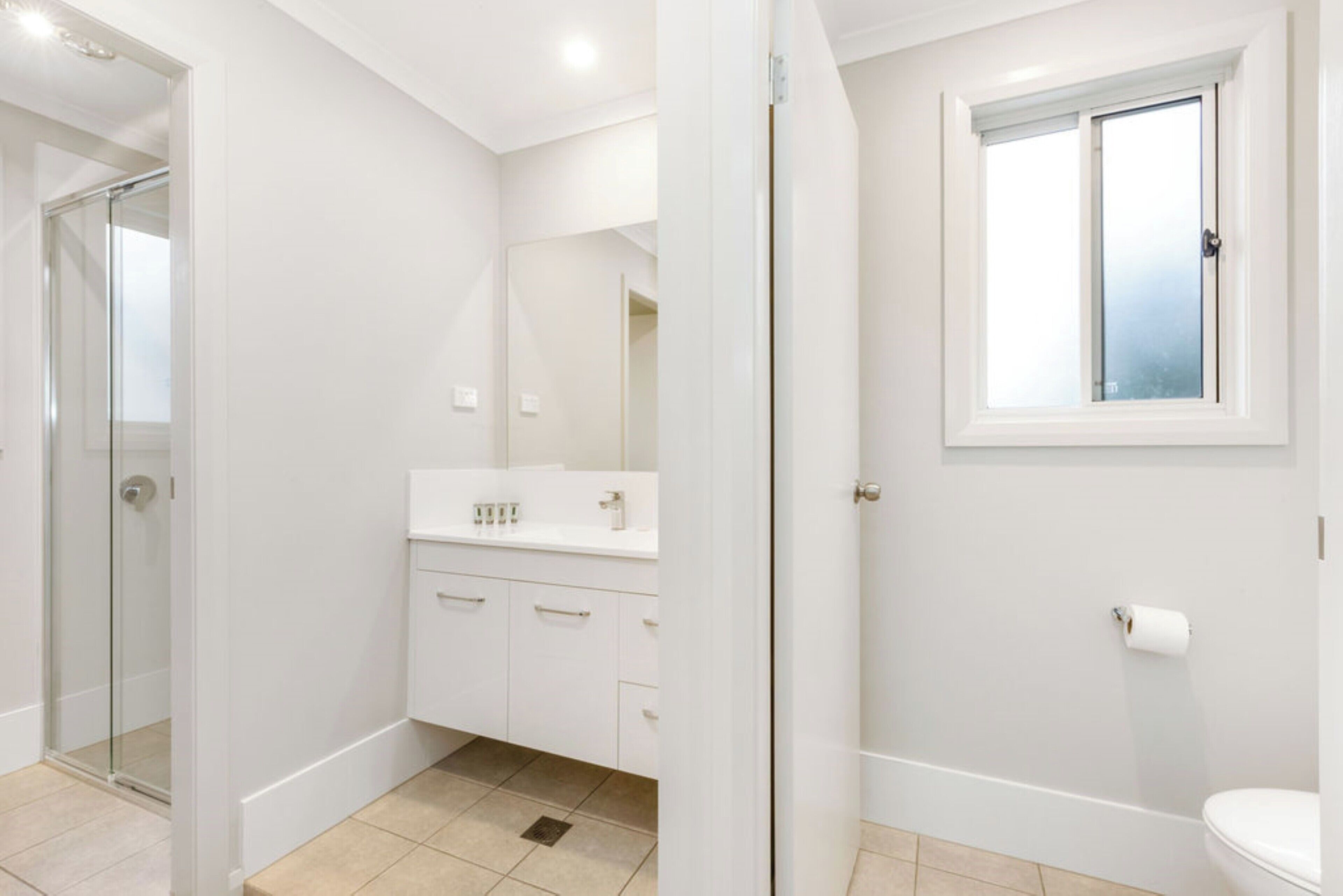 Un baño moderno con ducha de vidrio, armarios blancos, inodoro y ventana.