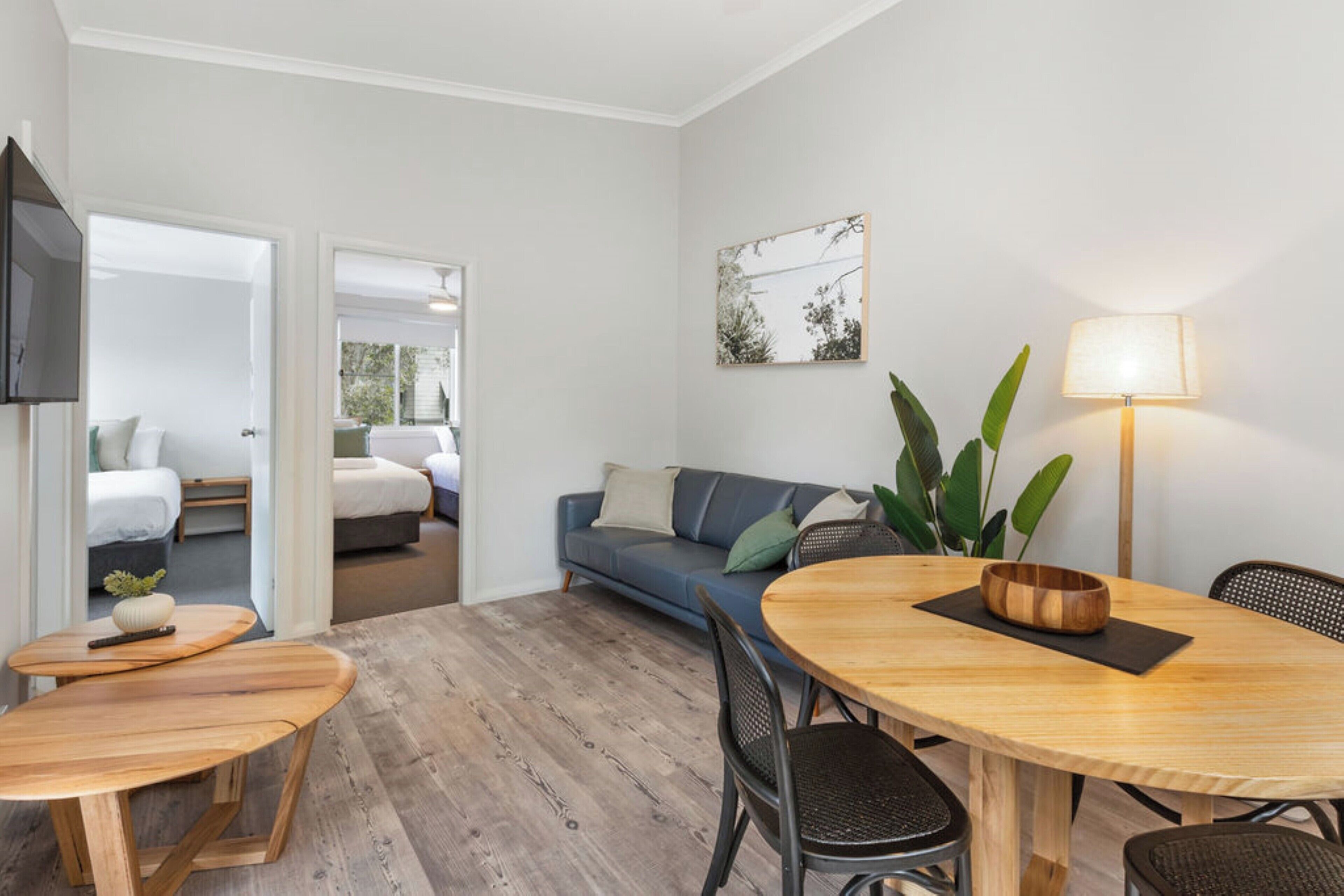 A modern hotel room with a wooden dining table, a sofa, and a TV mounted on the wall.