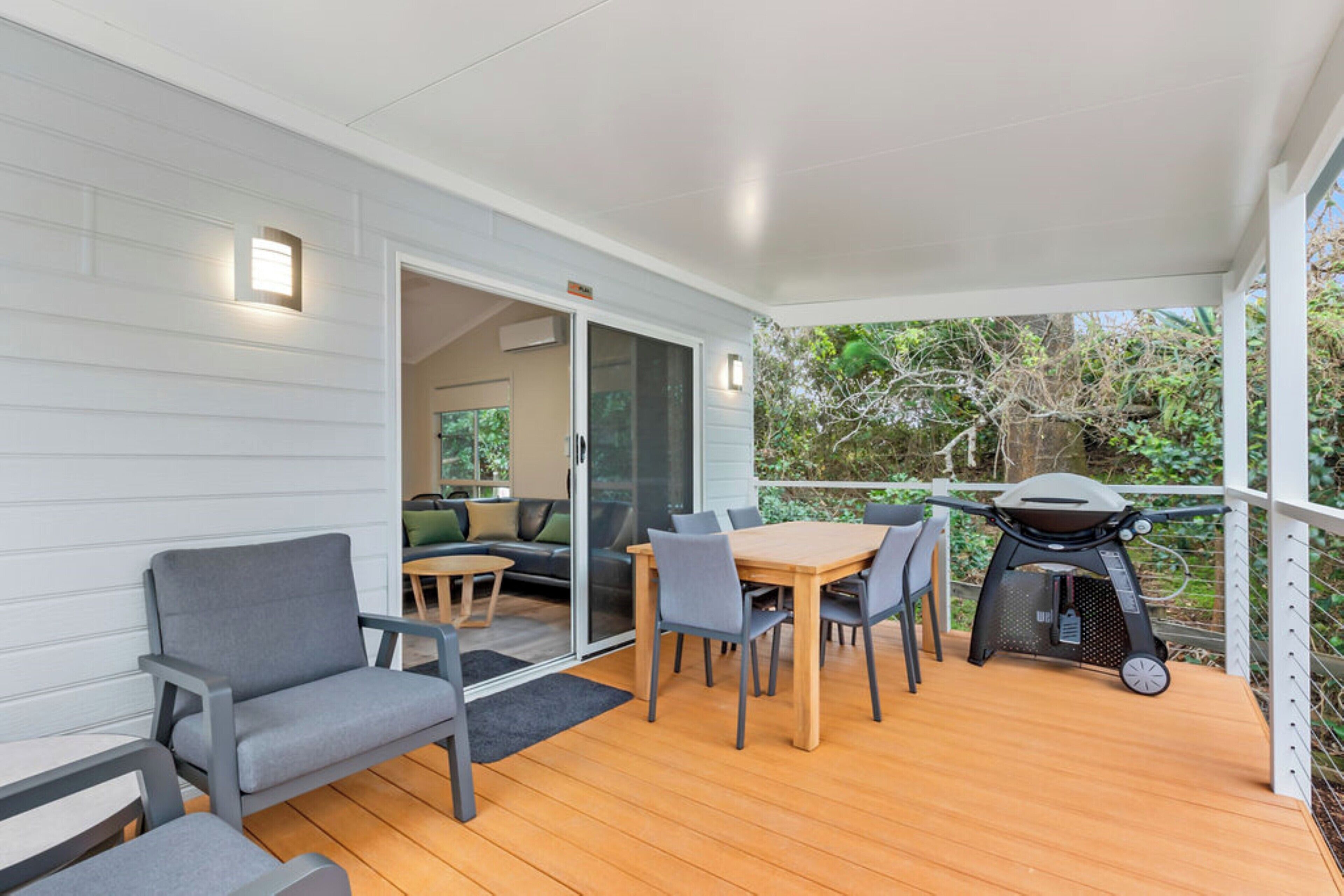 Une terrasse extérieure couverte, avec une table et des chaises en bois, un barbecue et une porte-fenêtre coulissante donnant sur un espace de détente intérieur.
