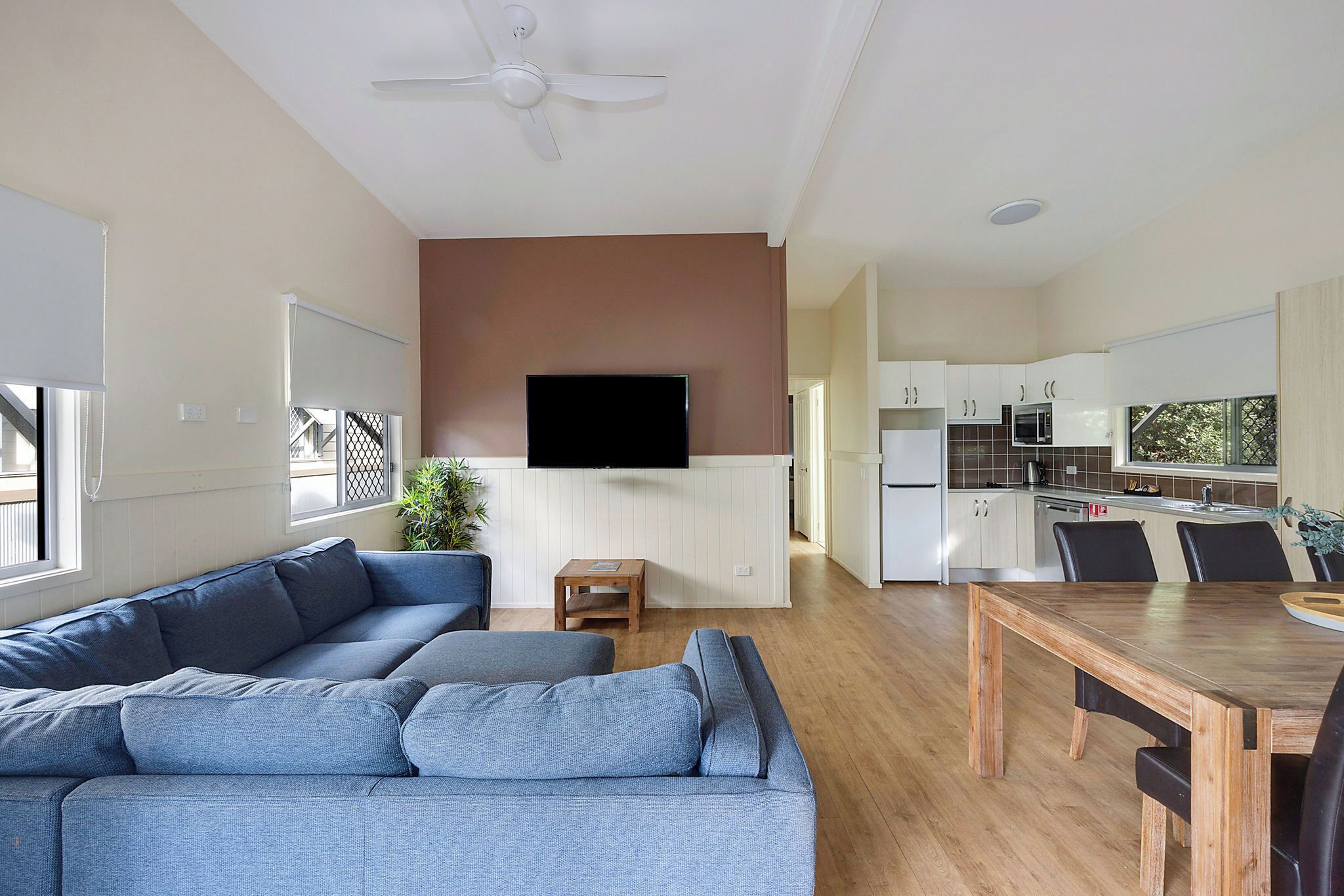 A modern living room with a grey sectional sofa, a wooden dining table, and a kitchen in the background.