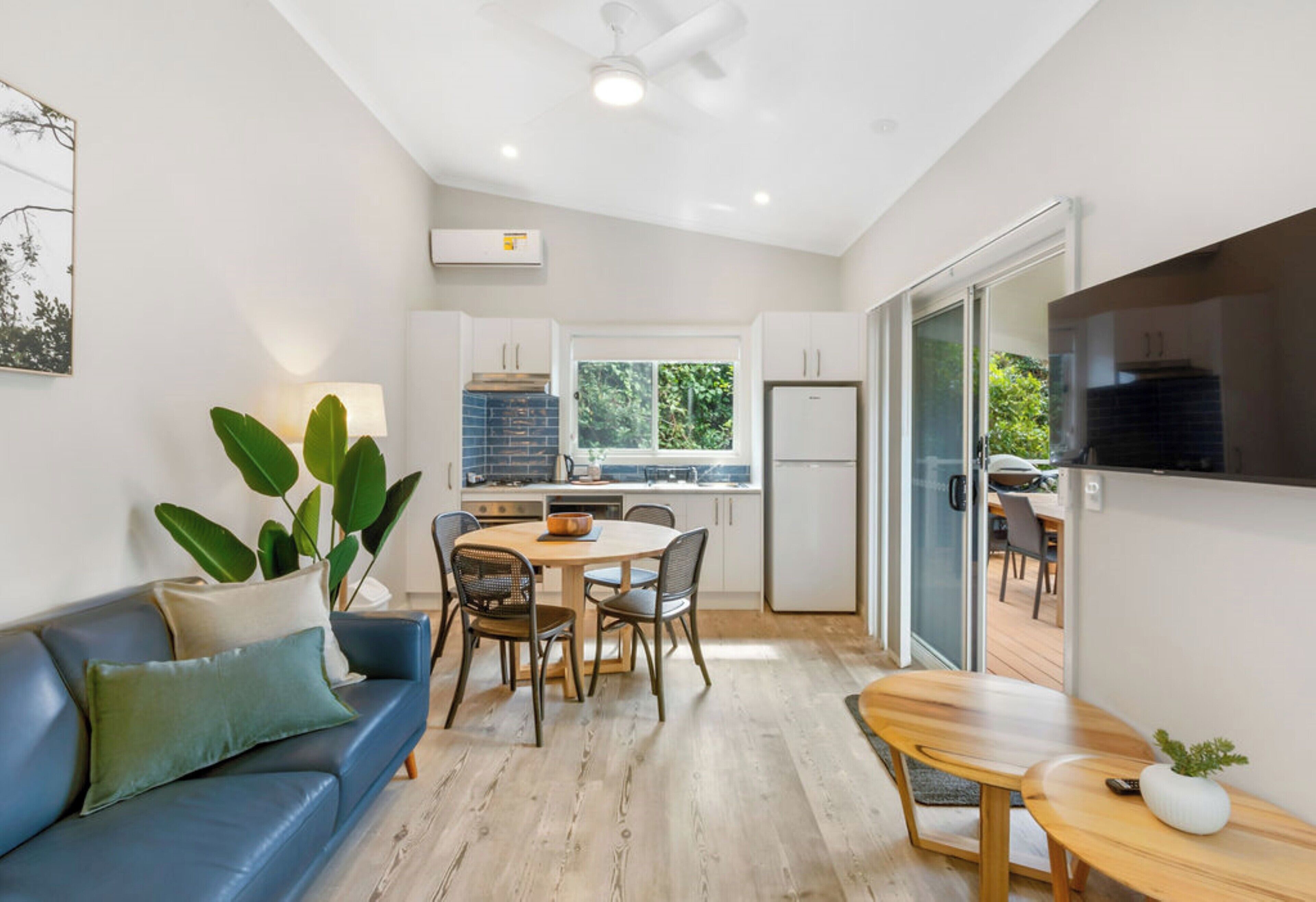 A modern kitchen with white cabinets, a dining area with a wooden table and chairs, and a blue sofa with cushions.