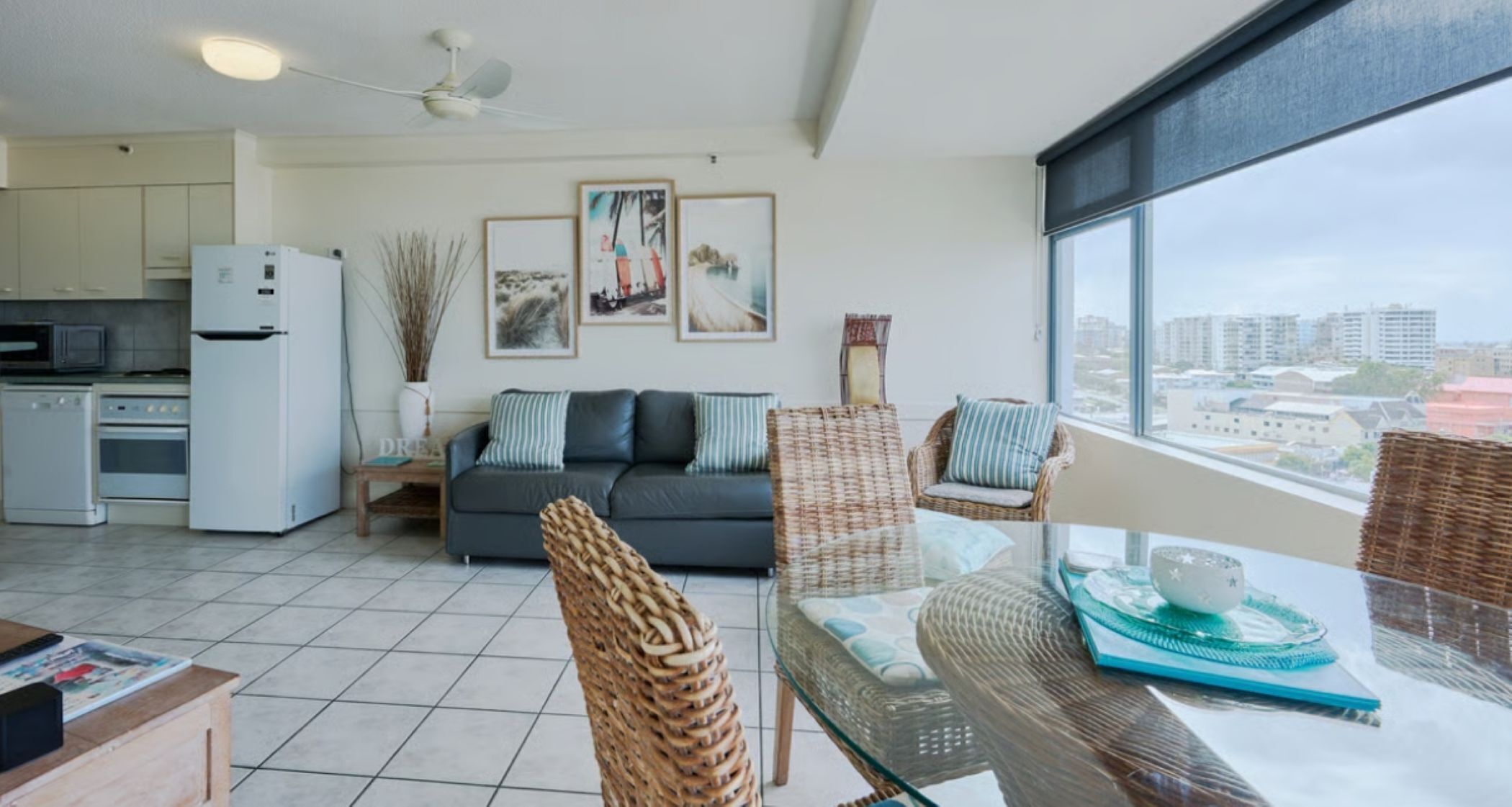 A modern living room with a glass dining table, wicker chairs, a grey sofa, and a city view through large windows.