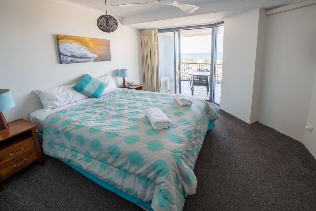 A hotel room with a large bed, a view of the sea through a sliding glass door, and a balcony with a table and chairs.