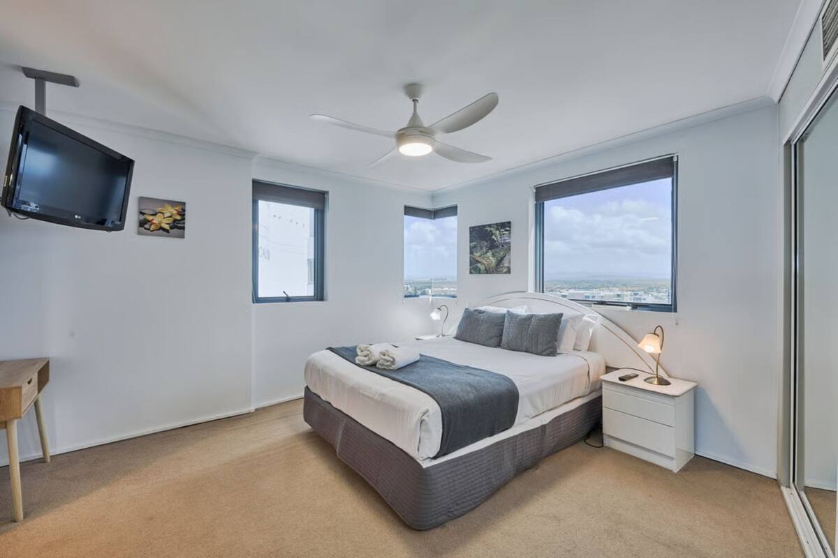 A modern bedroom with a bed, bedside tables, a ceiling fan, and large windows offering a city view.