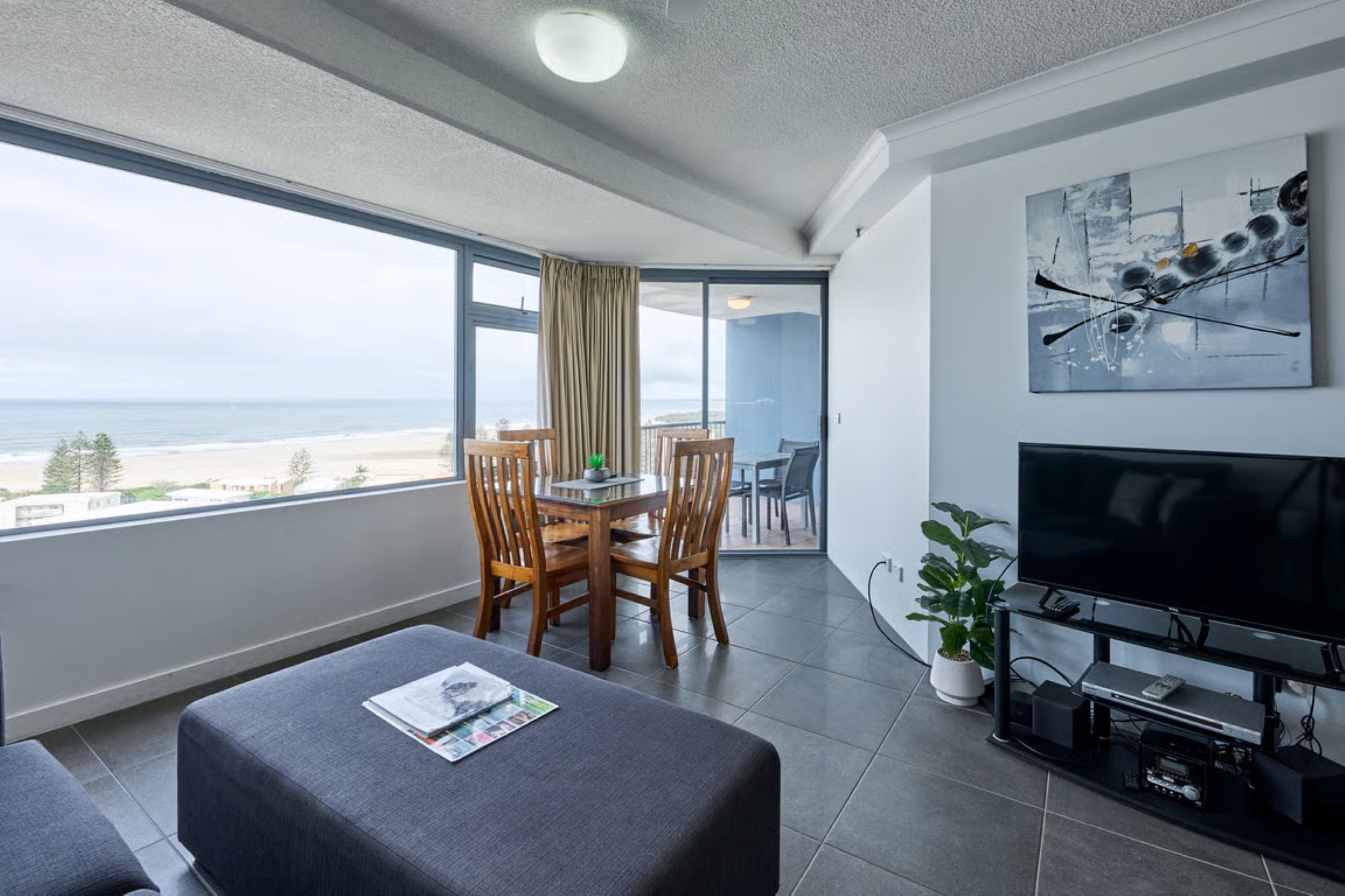 Una habitación moderna con balcón, una mesa de comedor con sillas, un televisor de pantalla plana y un gran ventanal con vistas a la playa.