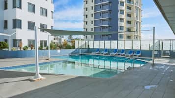 Une piscine entourée de chaises longues, elle-même bordée d’un immeuble à plusieurs étages doté de balcons.