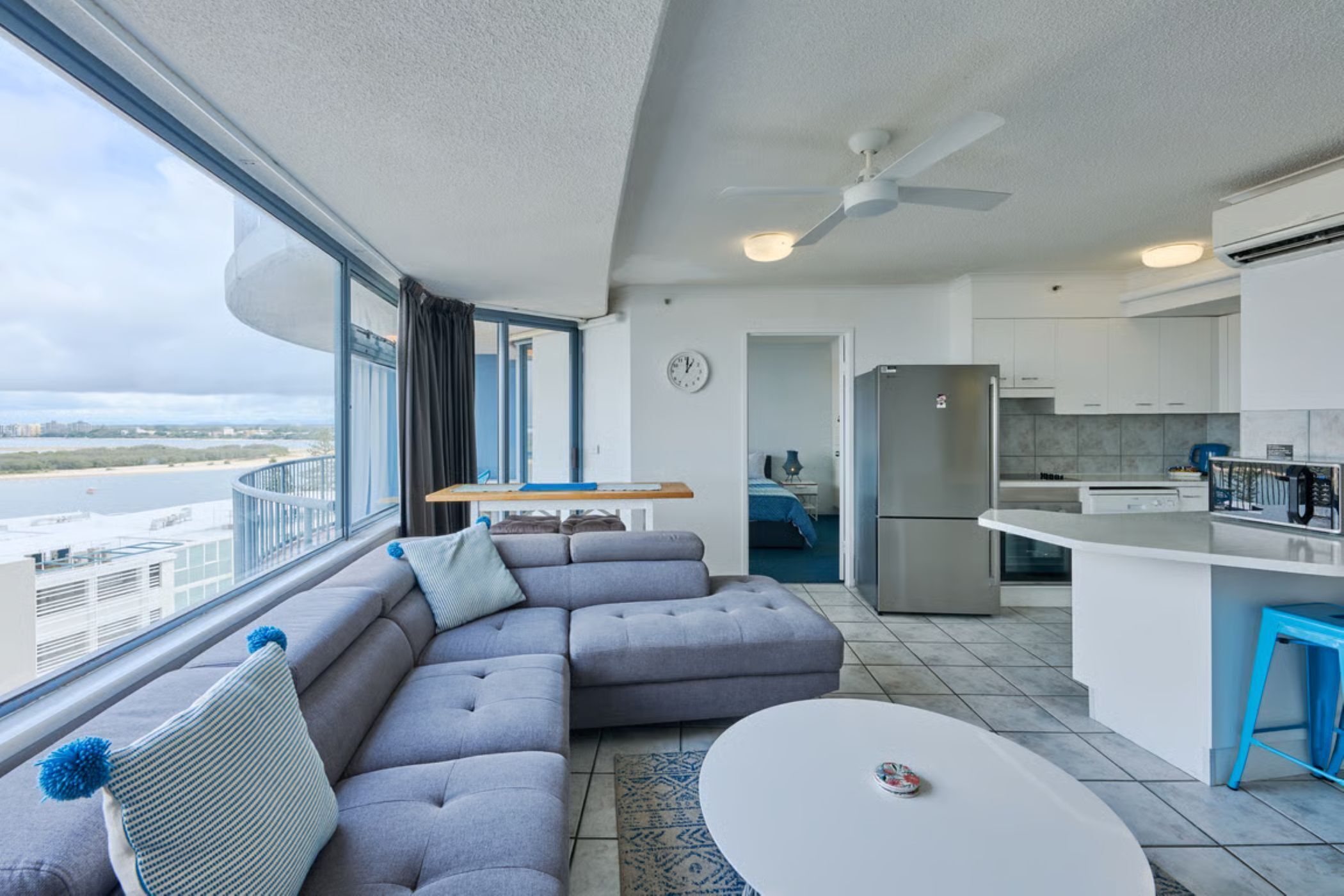 A modern living room with a grey sectional sofa, a round coffee table, and a view of a body of water through large windows.