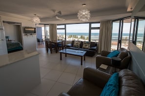 A modern living room with a sofa, coffee table, and dining area. A TV is mounted on the wall, and there are large windows offering a view of the ocean. - Centrepoint Holiday Apartments (Caloundra)