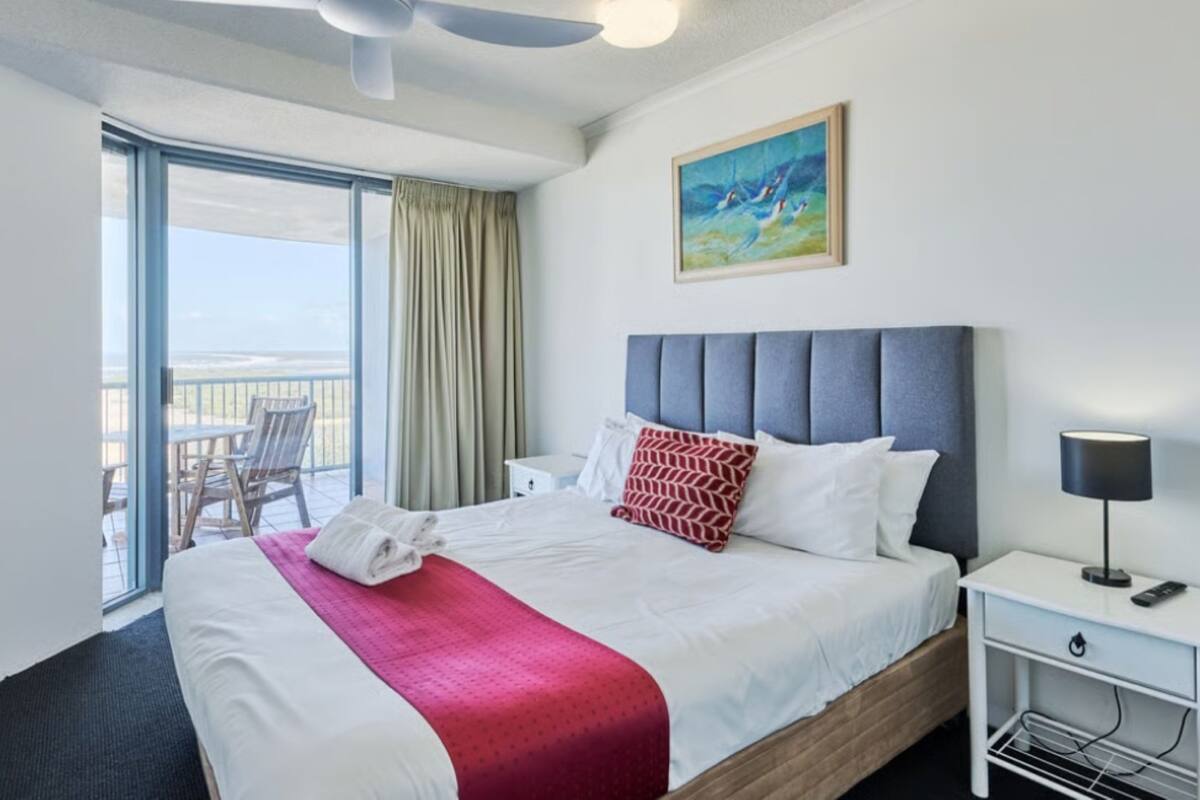 A hotel room with a bed, a nightstand, a lamp, a painting, and a view of the beach through the window.