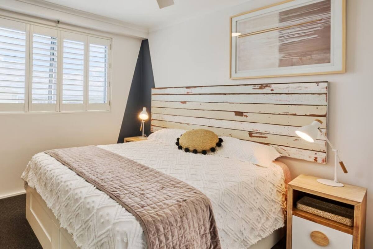 A bedroom with a bed, a nightstand, a lamp, a dresser, a window with blinds, and a framed picture on the wall.