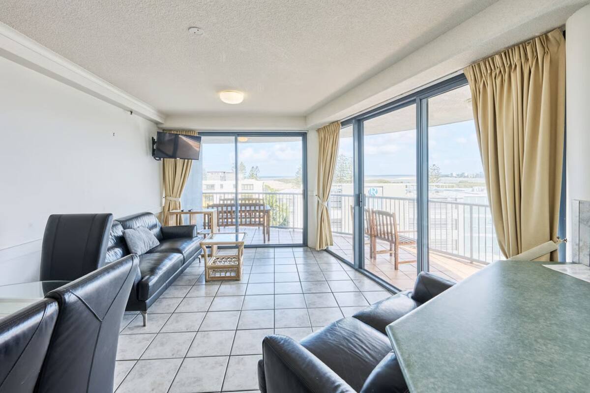 A modern living room with a sofa, a coffee table, and a dining table. There is a balcony with a view of the city.