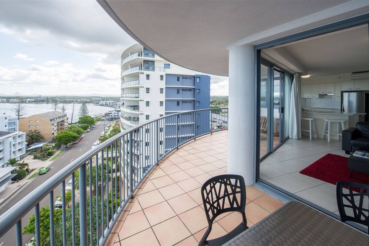 A balcony with a view of a river and buildings, a dining area with a red rug, and a kitchen with white cabinets.