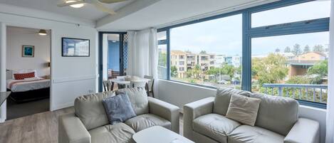A modern living room with two beige sofas, a white coffee table, and a ceiling fan. There is a balcony with a view of buildings and greenery.