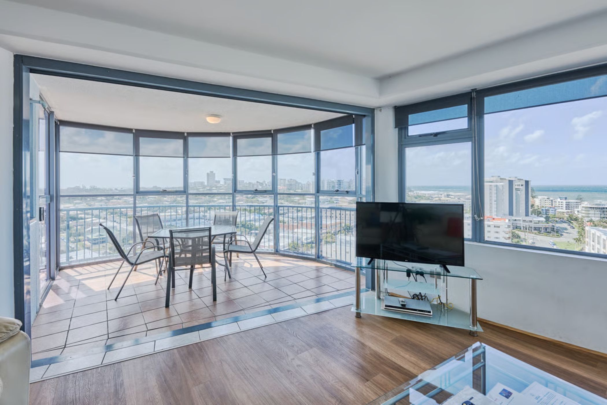 Uma varanda espaçosa com vista para a cidade, uma mesa de vidro com cadeiras e uma TV de tela plana em um suporte de vidro.