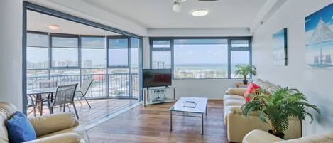 A spacious living room with a balcony, large windows, a ceiling fan, and a flat-screen TV.