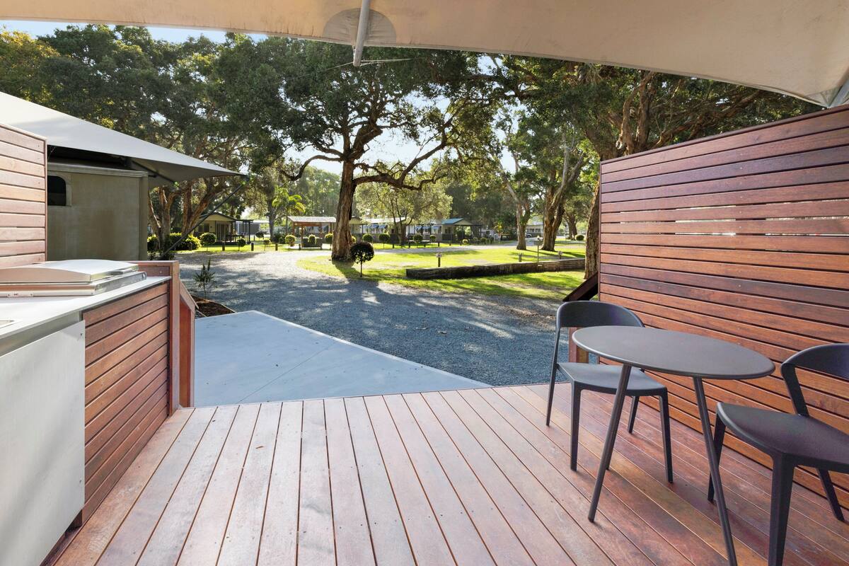 A wooden deck with a barbecue area, outdoor seating, and a view of a park with trees and a building in the background.