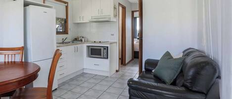 Una cocina compacta con armarios blancos, microondas y refrigerador. Hay una mesa de comedor con sillas y un sofá de cuero negro con cojines.