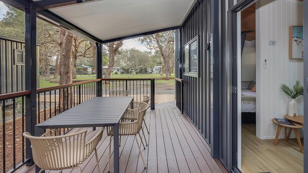 Un espacio al aire libre techado con una mesa y sillas, vistas a los árboles y casas circundantes, y una puerta que da a una habitación con una cama y una mesita.