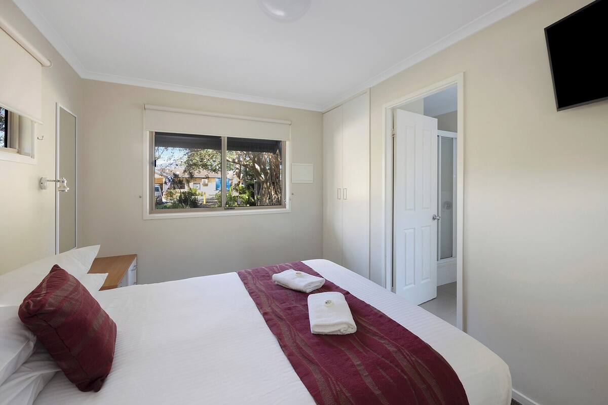 A hotel room with a bed, a window, a door, and a TV on the wall.