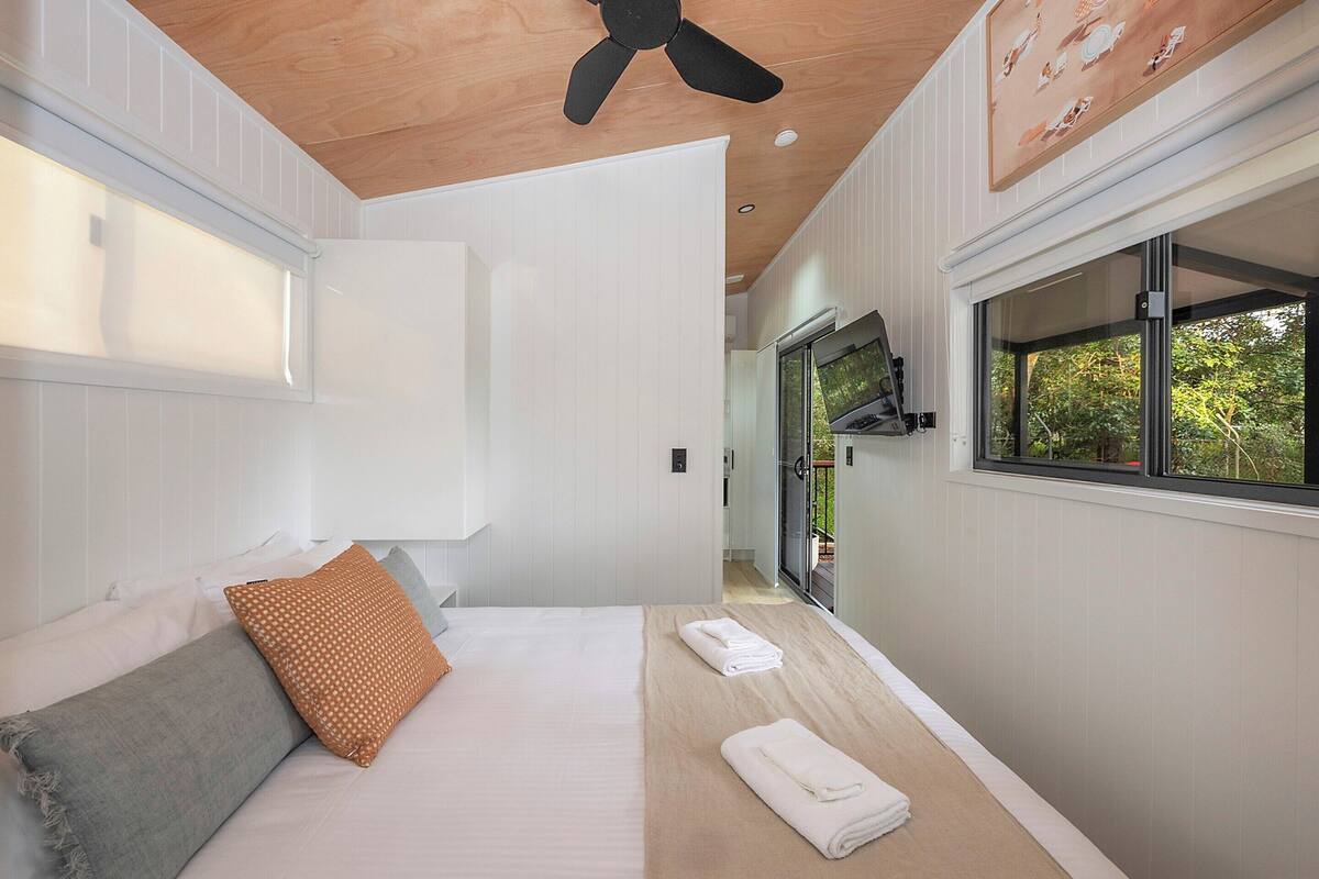 A bedroom with a bed, pillows, a ceiling fan, and a window with a view of greenery.
