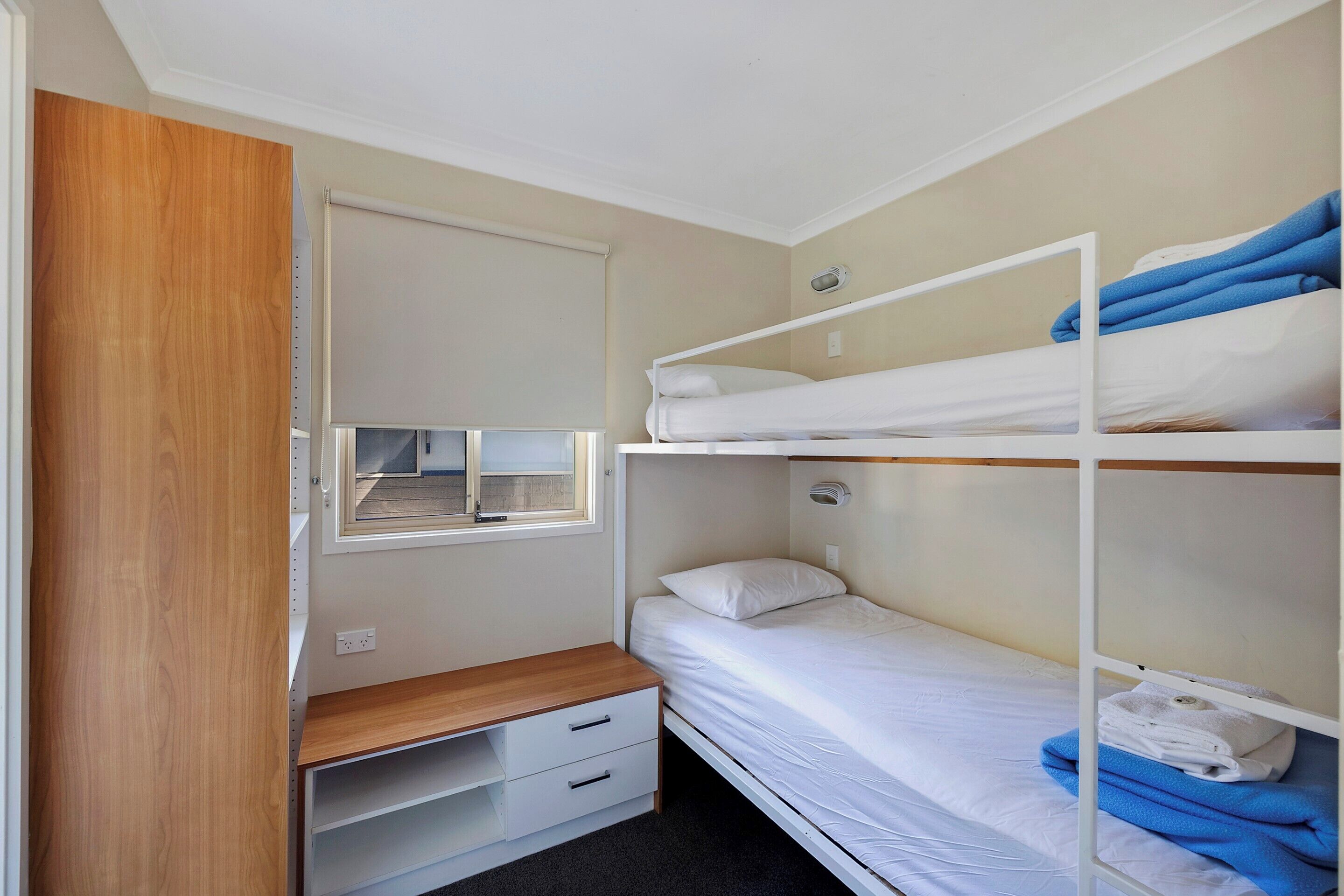 Een kleine kamer met een stapelbed, een klein bureau met lades, een raam en een houten garderobe.