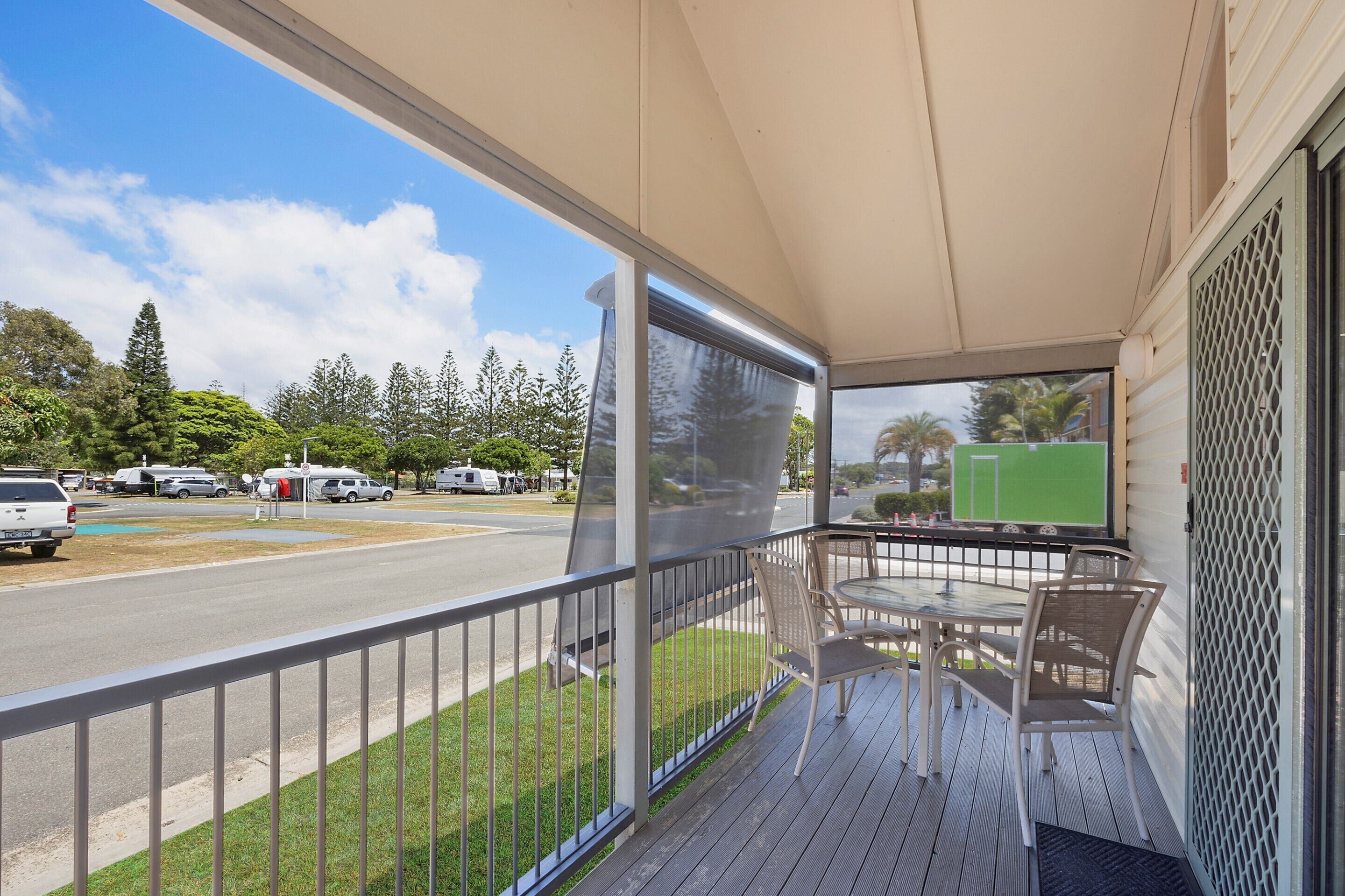 A covered outdoor area with a table and chairs, a view of a road with parked vehicles, and greenery in the background.