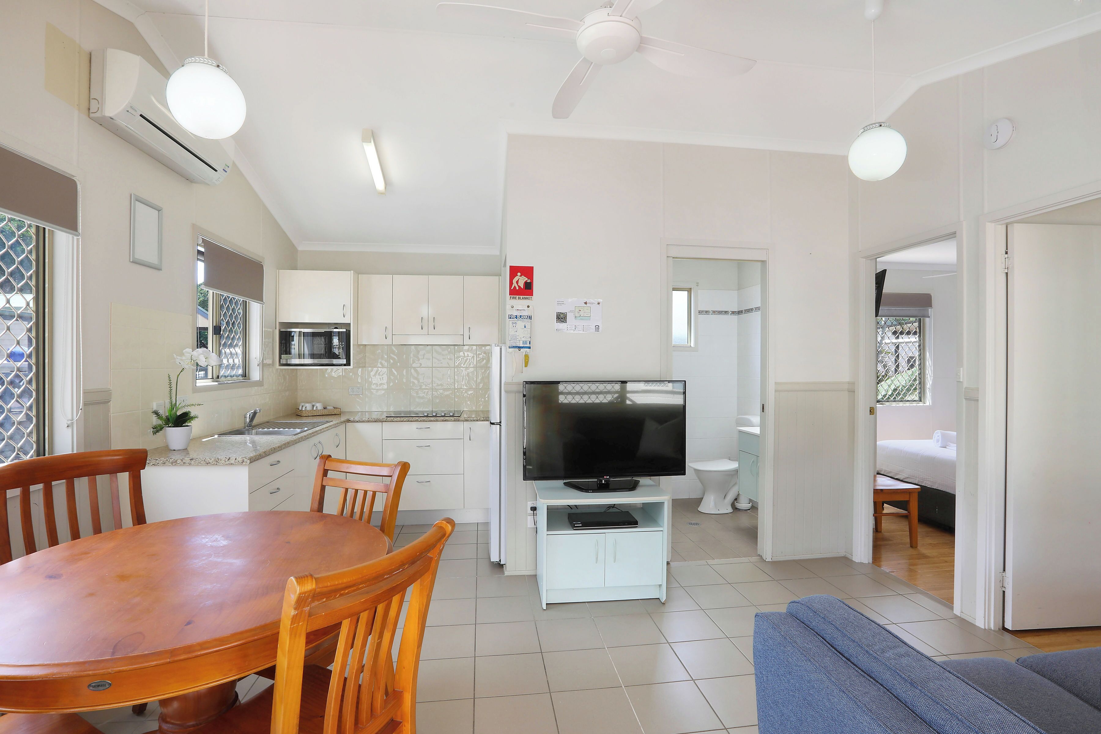 A modern kitchen with white cabinets, a dining table, and chairs. There is a TV on a stand, a ceiling fan, and a window with a view.