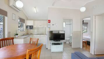 A modern kitchen with white cabinets, a dining table, and chairs. There is a TV on a stand, a ceiling fan, and a window with a view.