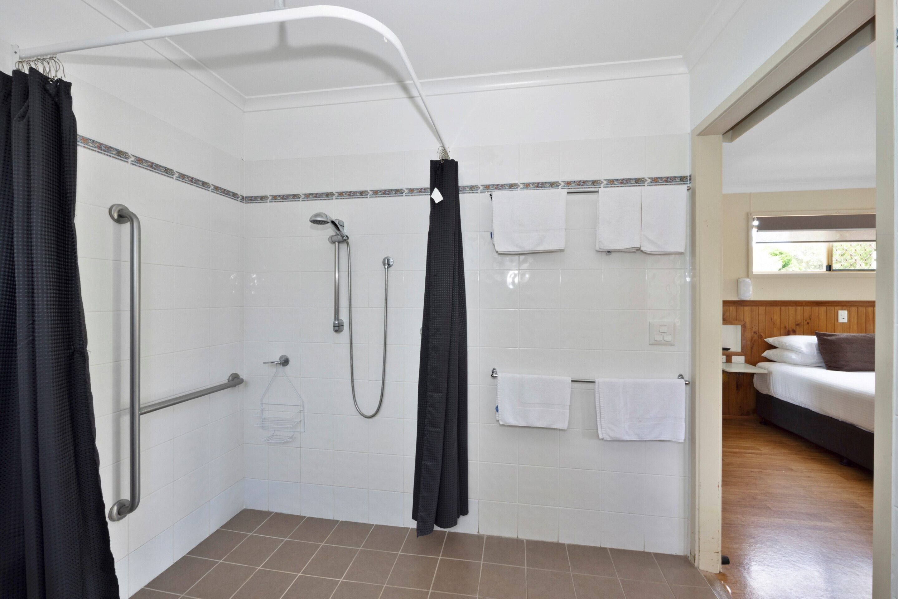 A bathroom with white tiled walls, a shower with a handheld showerhead, a grab bar, and a towel rack with multiple towels.