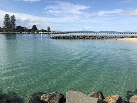A clear coastal view with a rocky shoreline, a pier extending into the water, and a distant mountain range.