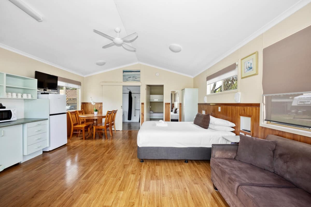 A modern hotel room with a wooden floor, a large bed, a sofa, a dining area with chairs, a TV, and a ceiling fan.