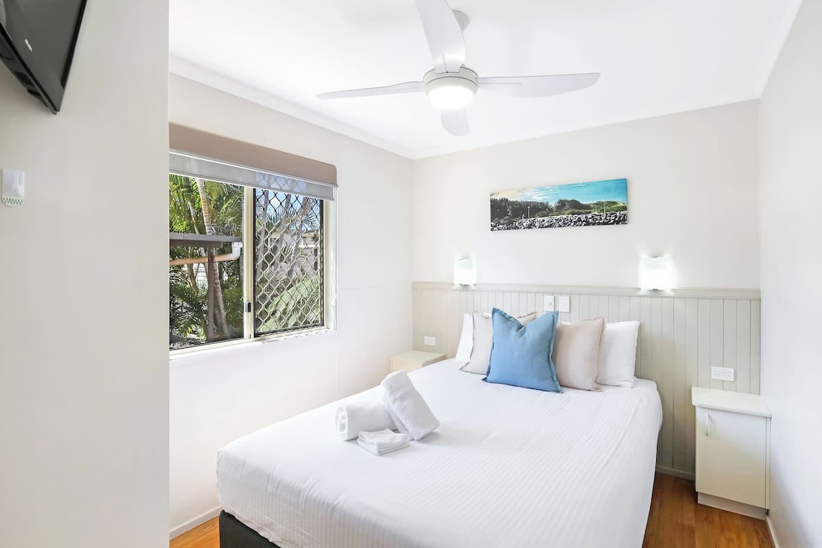 A neatly made bed with white linens and a blue pillow, a ceiling fan, a window with a view of palm trees, and a wall-mounted picture.