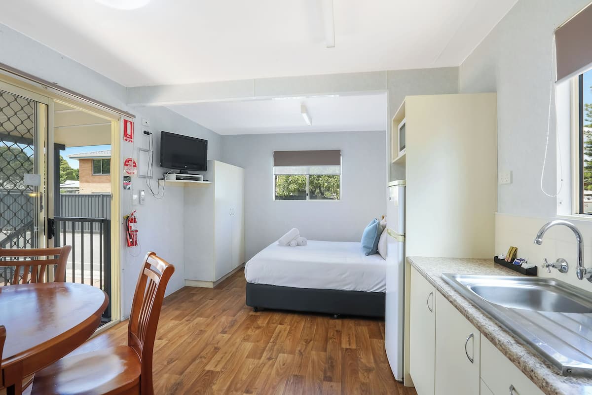 A compact room with a bed, a dining table, chairs, a TV, a window, and a kitchenette.