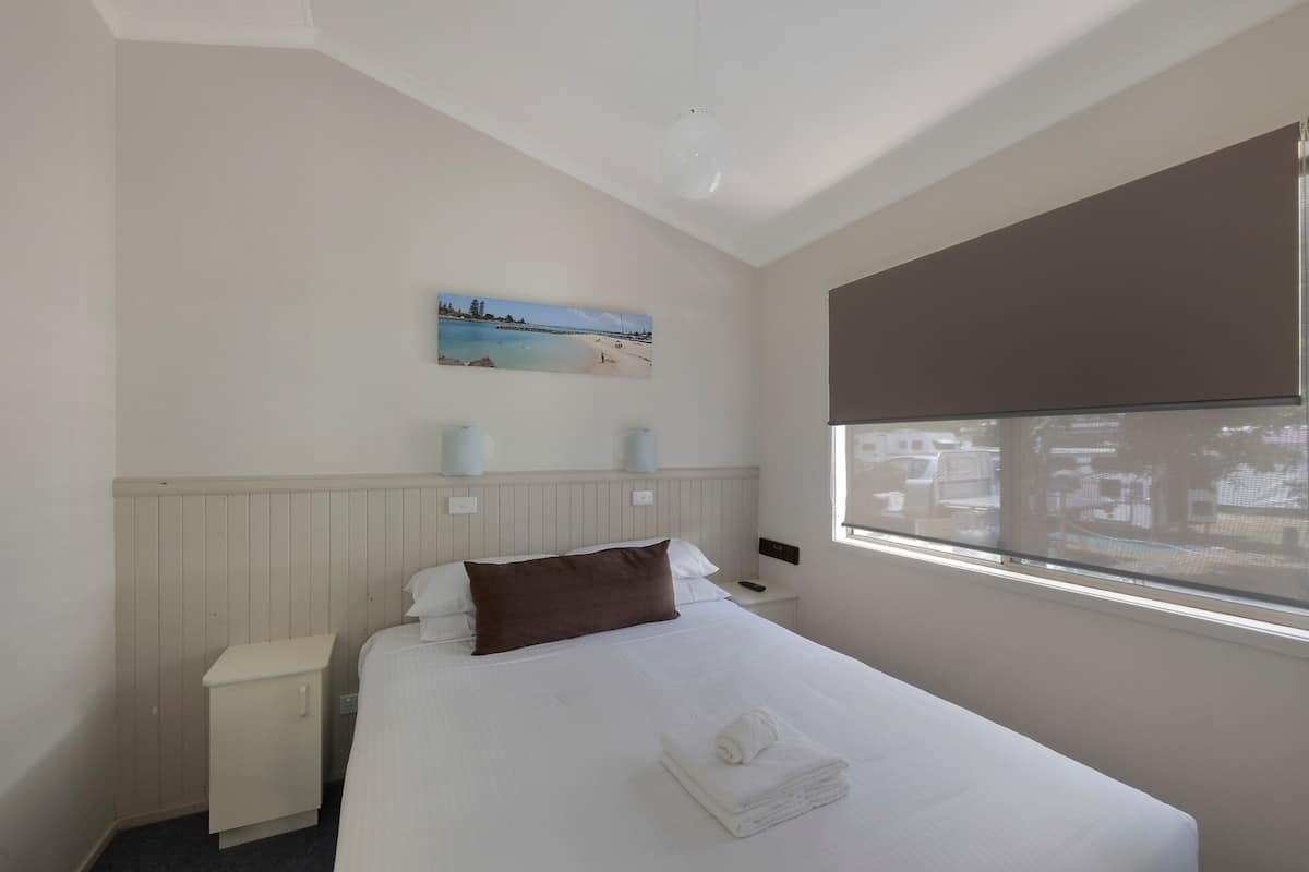 A hotel room with a bed, a nightstand, a wall-mounted lamp, a window with a view, and a painting on the wall.