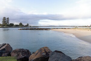 A coastal beach with a rocky breakwater, calm water, and a distant bridge.