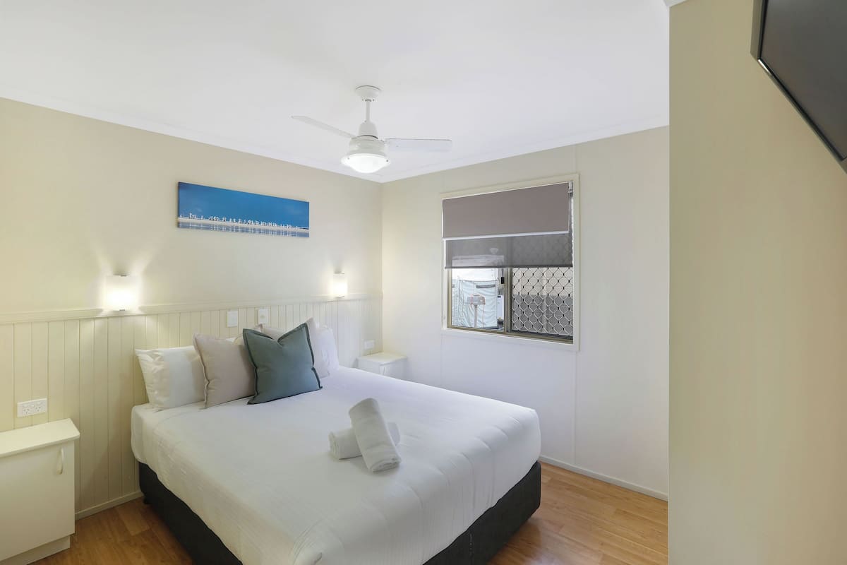 A neatly made bed with white linens and a folded towel, a ceiling fan, a window with a view, and a wall-mounted artwork.