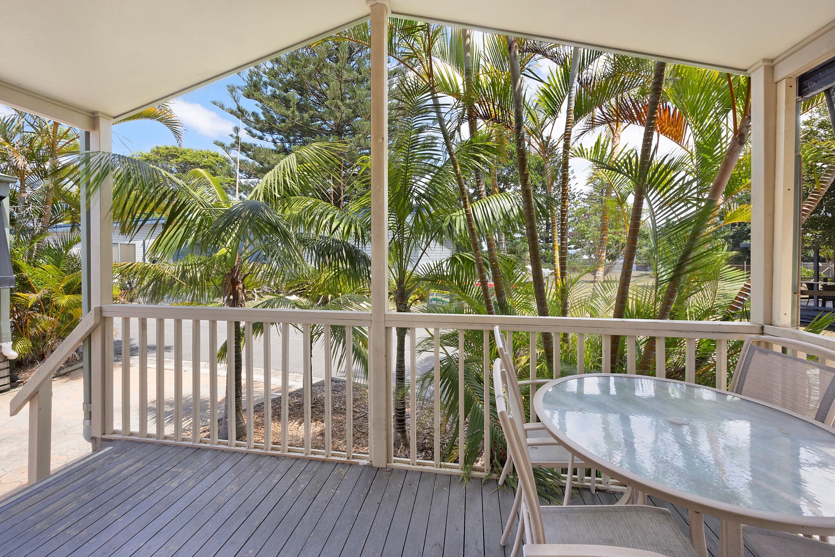 A covered outdoor area with a glass table and chairs, surrounded by a white railing and palm trees.