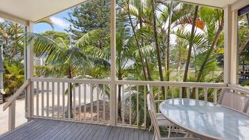 A covered outdoor area with a glass table and chairs, surrounded by a white railing and palm trees.