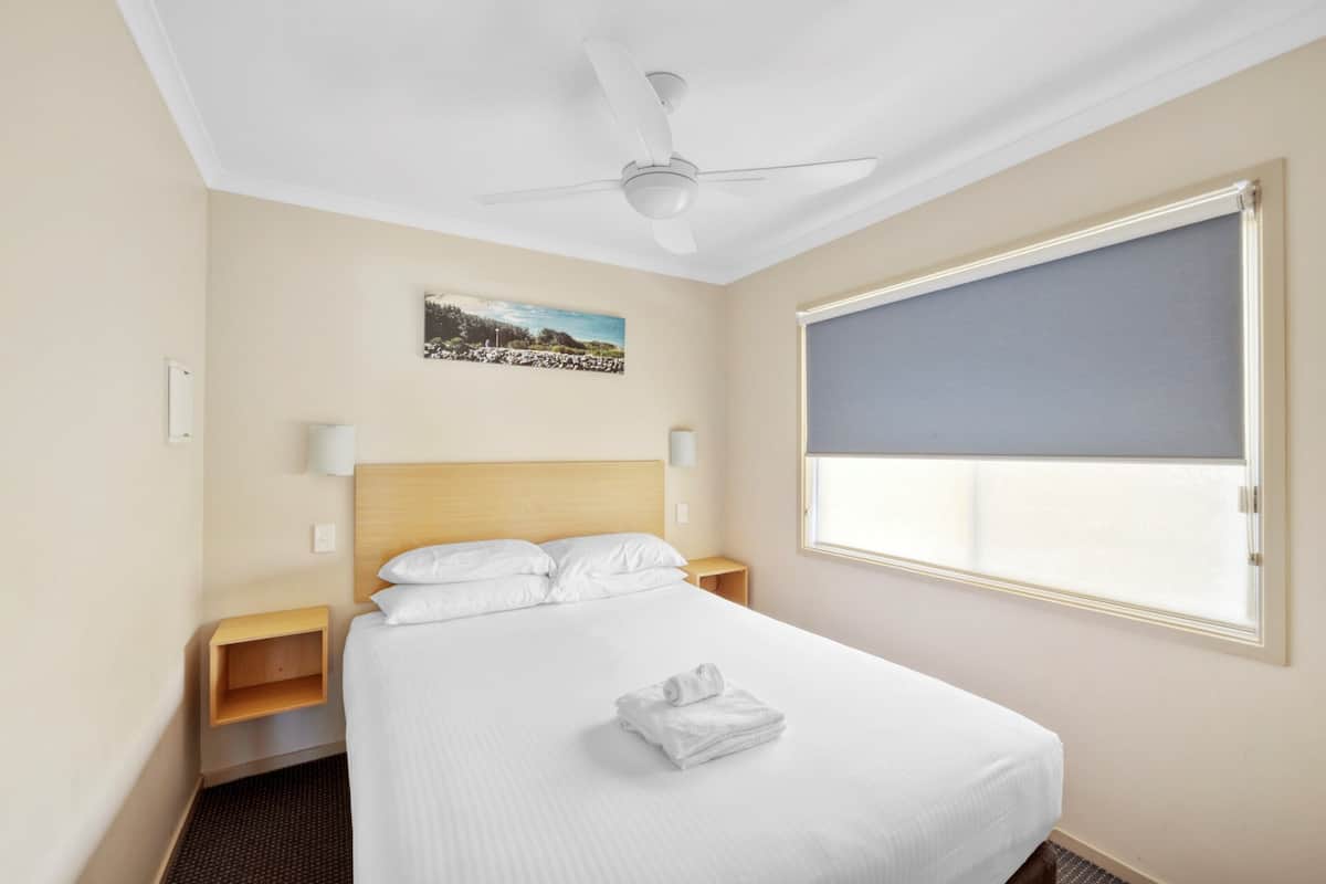 A hotel room with a bed, a ceiling fan, a window with blinds, a nightstand, and a wall-mounted picture.