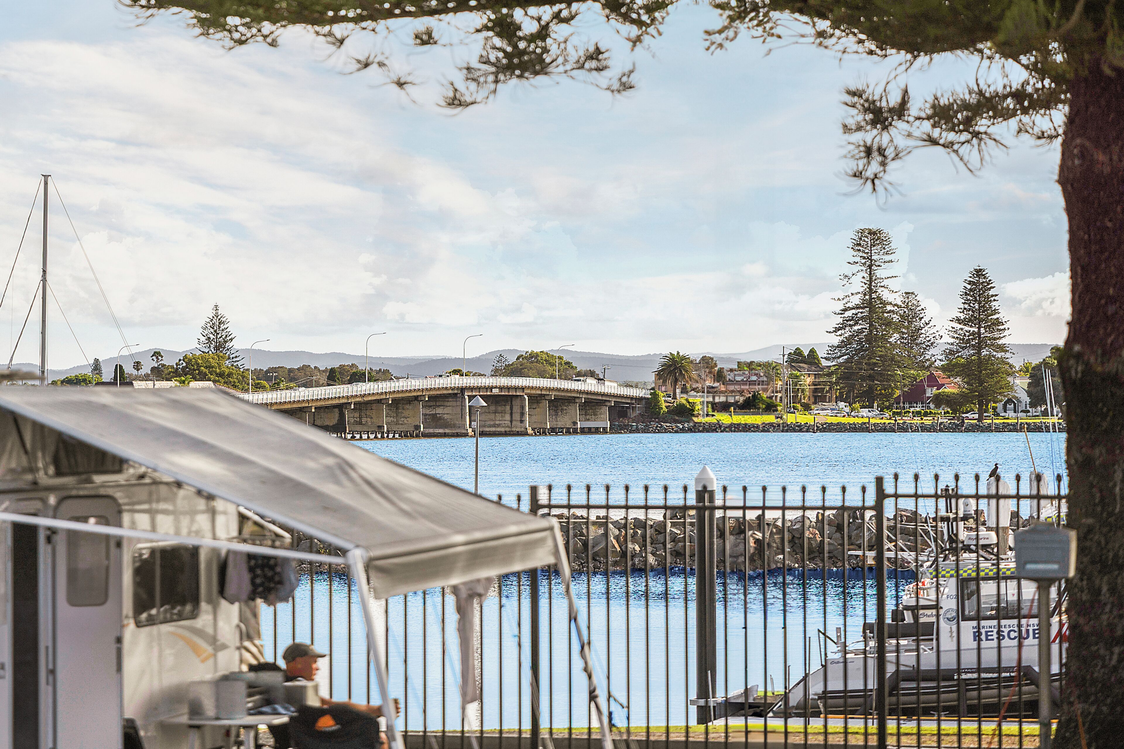 A waterfront view with a bridge, a boat, and a fenced area with a tent.