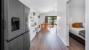 A modern kitchen with a built-in refrigerator, wooden dining table, and a sliding door leading to a bedroom with a bed and a view of the outdoors. - Reflections Forster Beach - Holiday Park (Forster)