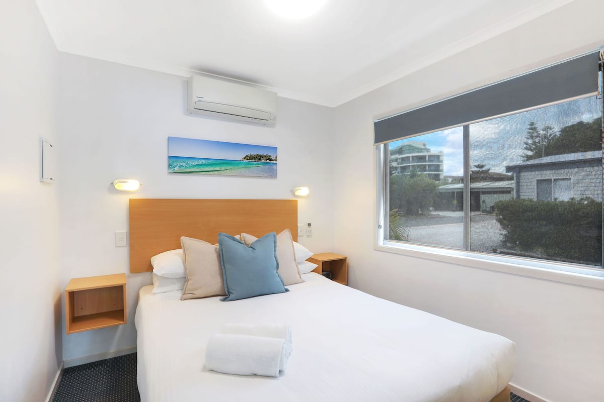 Una cama bien hecha con edredón blanco y almohada azul, una mesita de noche de madera, aire acondicionado instalado en la pared, un cuadro enmarco con una playa, y un ventanal con vistas a edificios y vegetación.