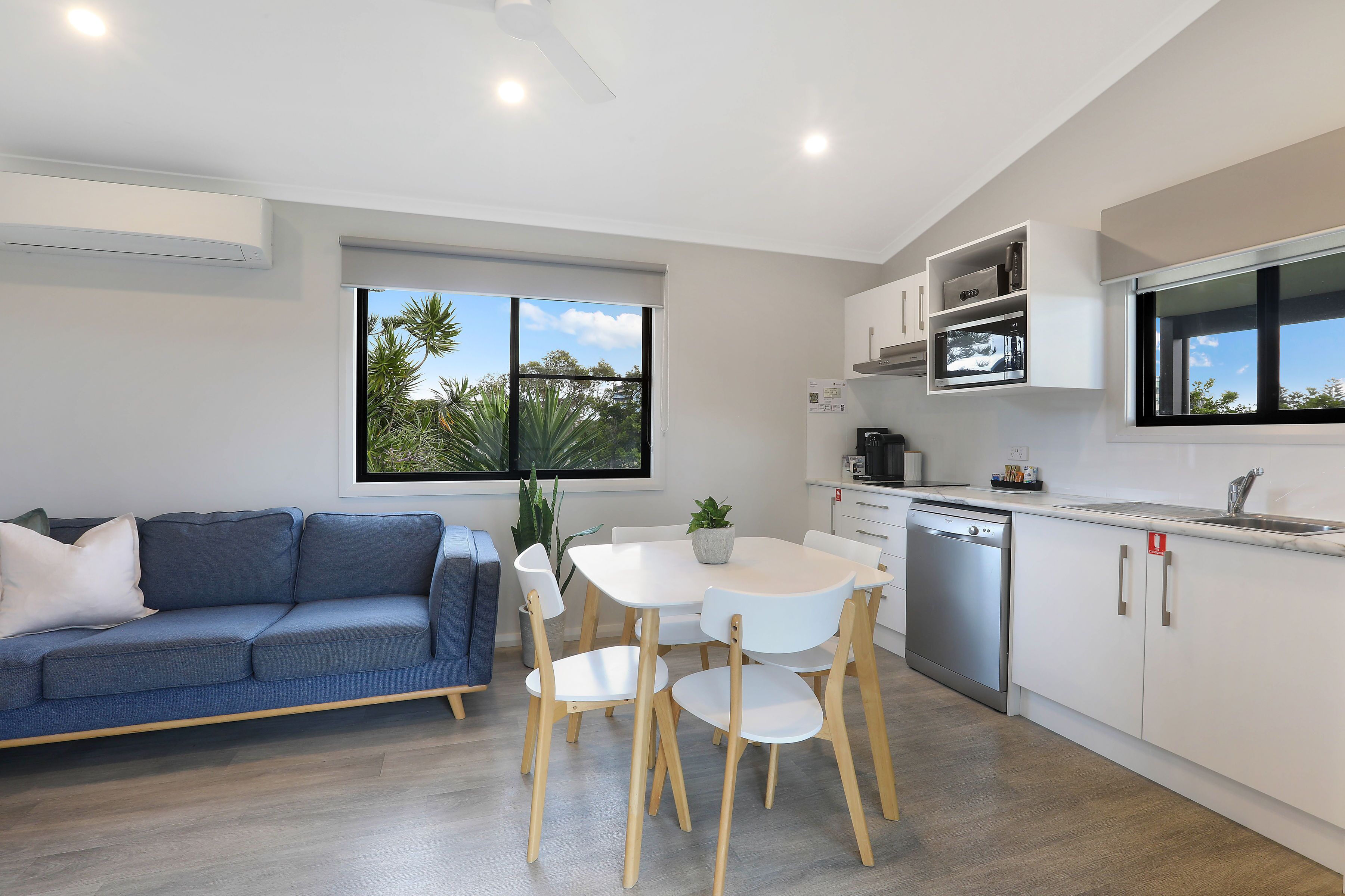 A modern kitchen with white cabinets, a built-in oven, and a dishwasher. There is a dining table with chairs and a blue sofa in the living area.