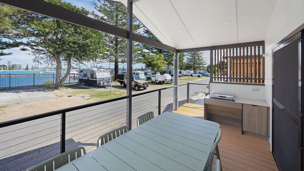 A covered outdoor area with a table and chairs, a small kitchen area, and a view of a parking lot with RVs and a body of water. - Reflections Forster Beach - Holiday Park (Forster)