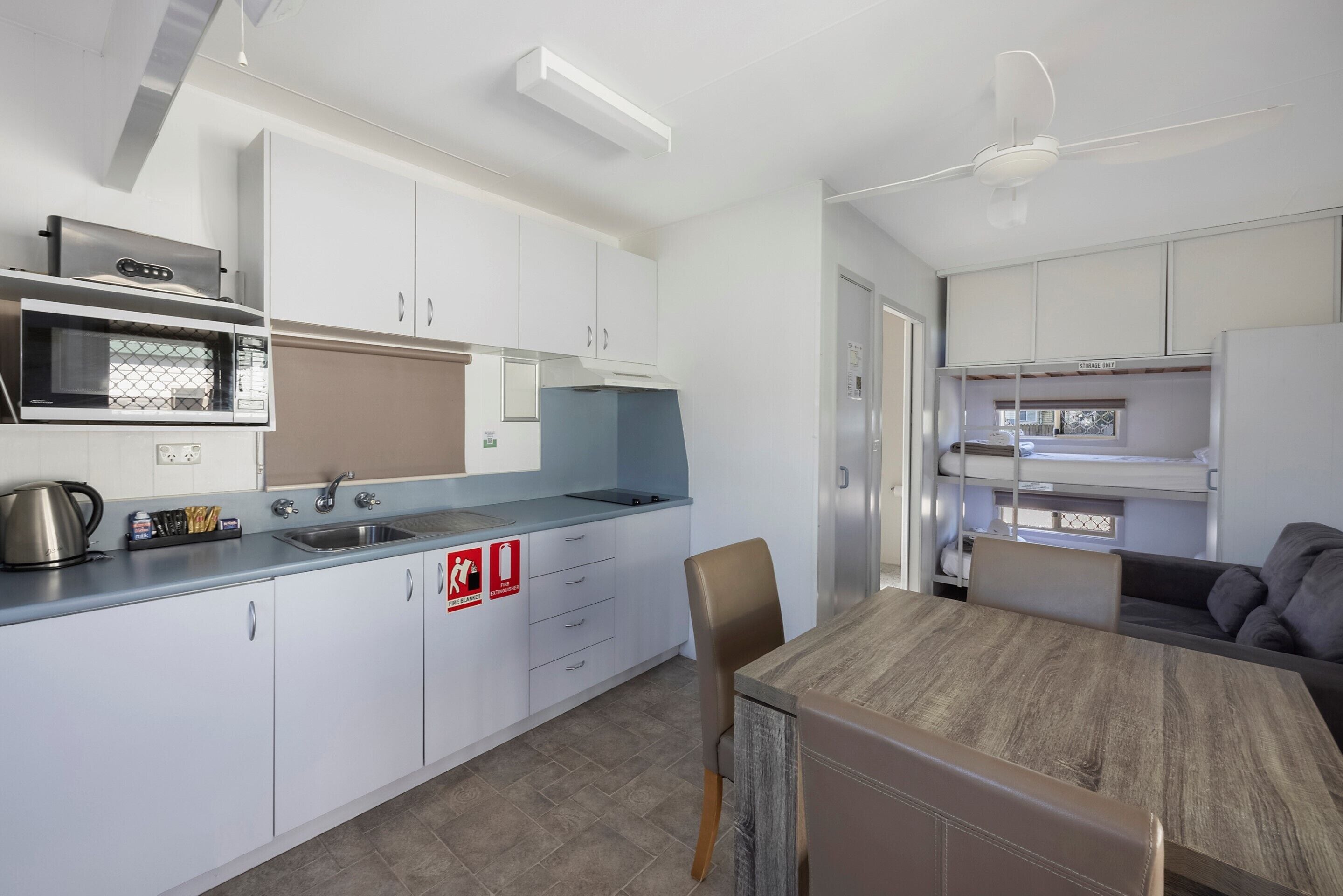 A modern kitchen with white cabinets, a stainless steel sink, a microwave, and a kettle. There is a dining table with chairs and a ceiling fan.