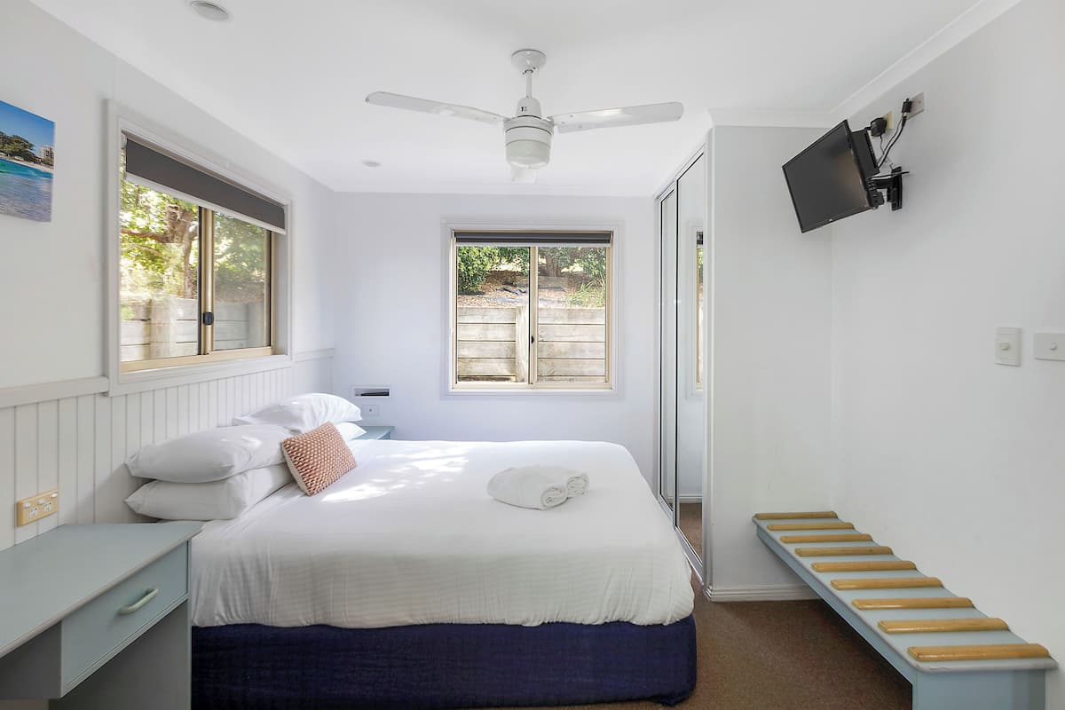 Un dormitorio con cama, escritorio, ventilador de techo, televisor montado en la pared y ventana.