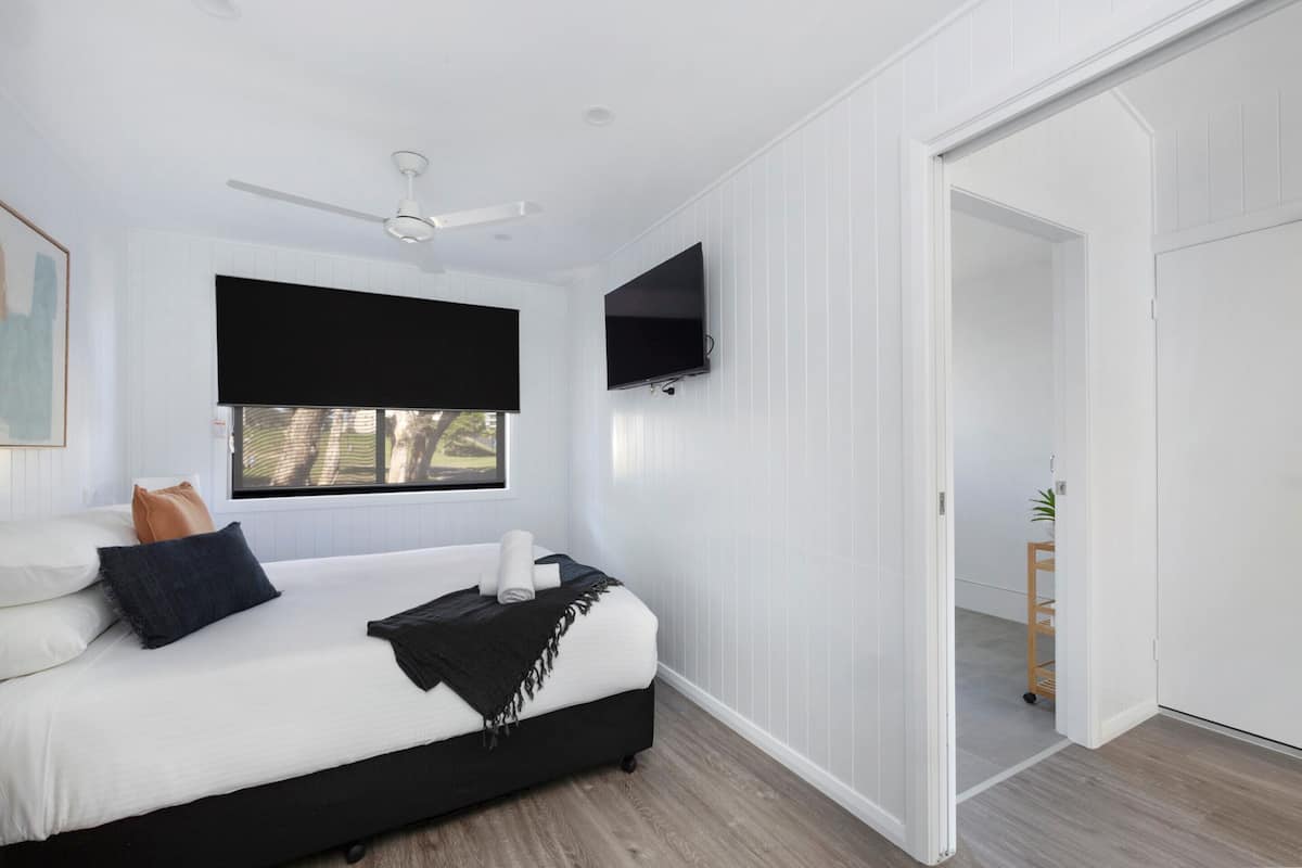 Un dormitorio con cama, ventilador de techo, televisor montado en la pared y ventana con persianas.