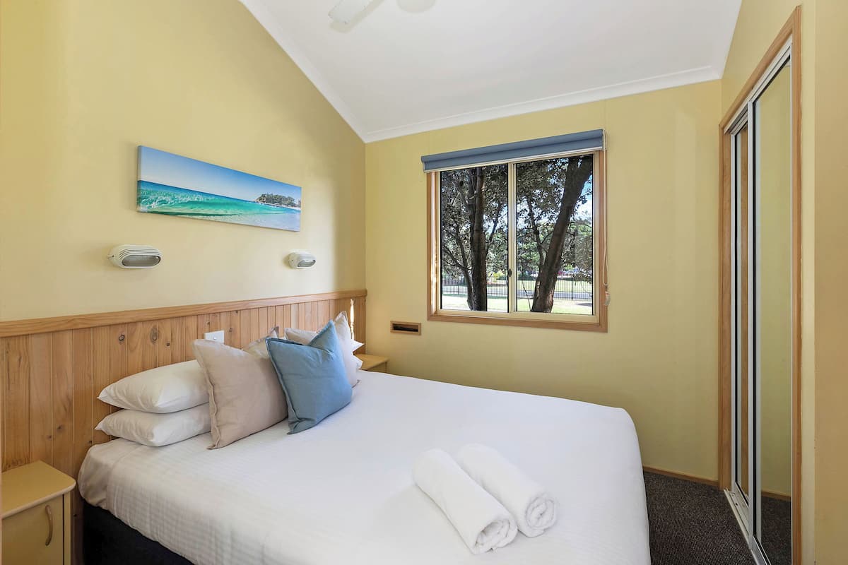 Una habitación de hotel con cama, almohadas, un cuadro, una ventana y vistas a árboles.