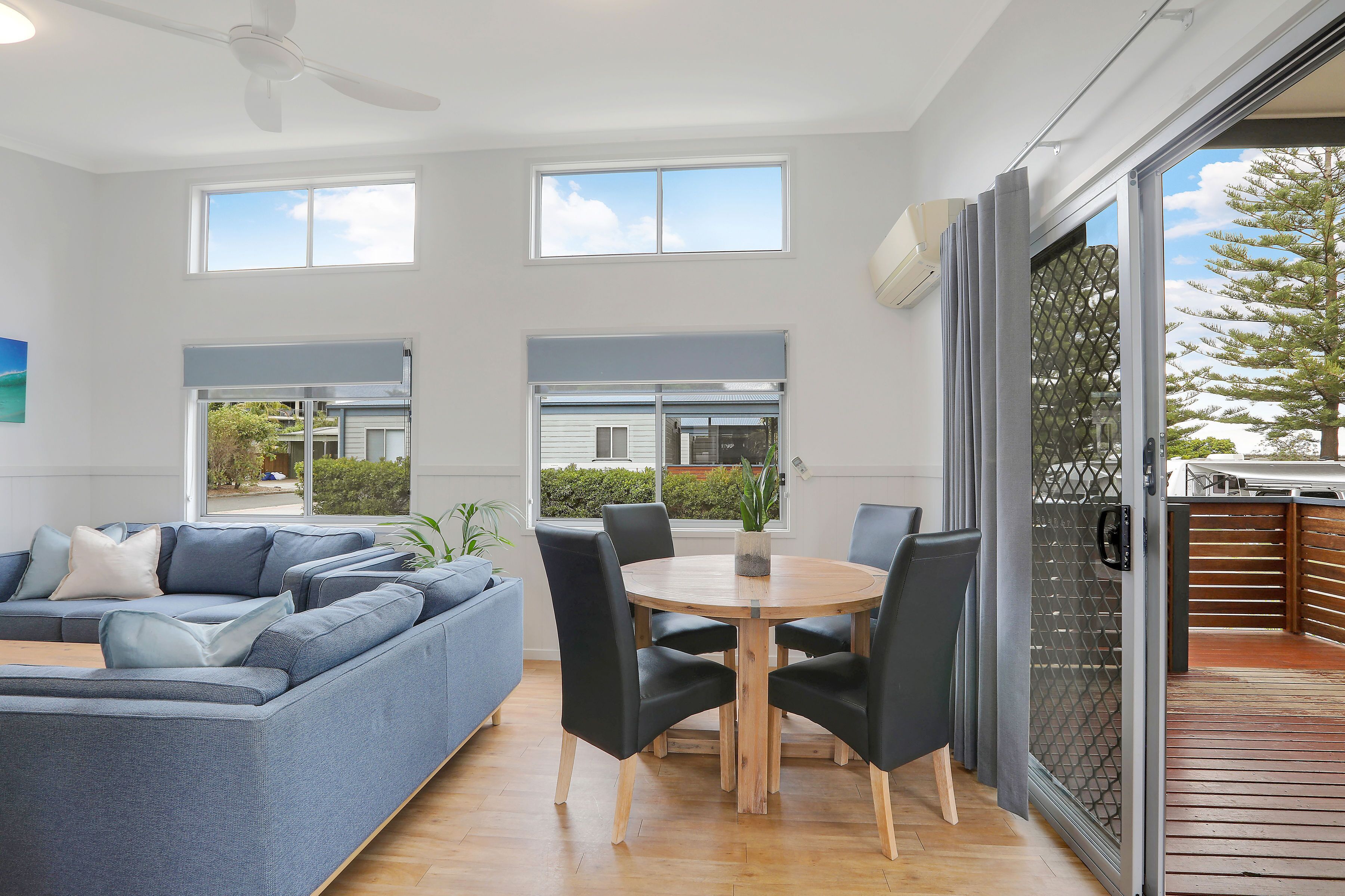 Una sala de estar moderna con un sofá seccional gris, una mesa de comedor de madera con sillas y ventanas amplias que ofrecen vistas al exterior.