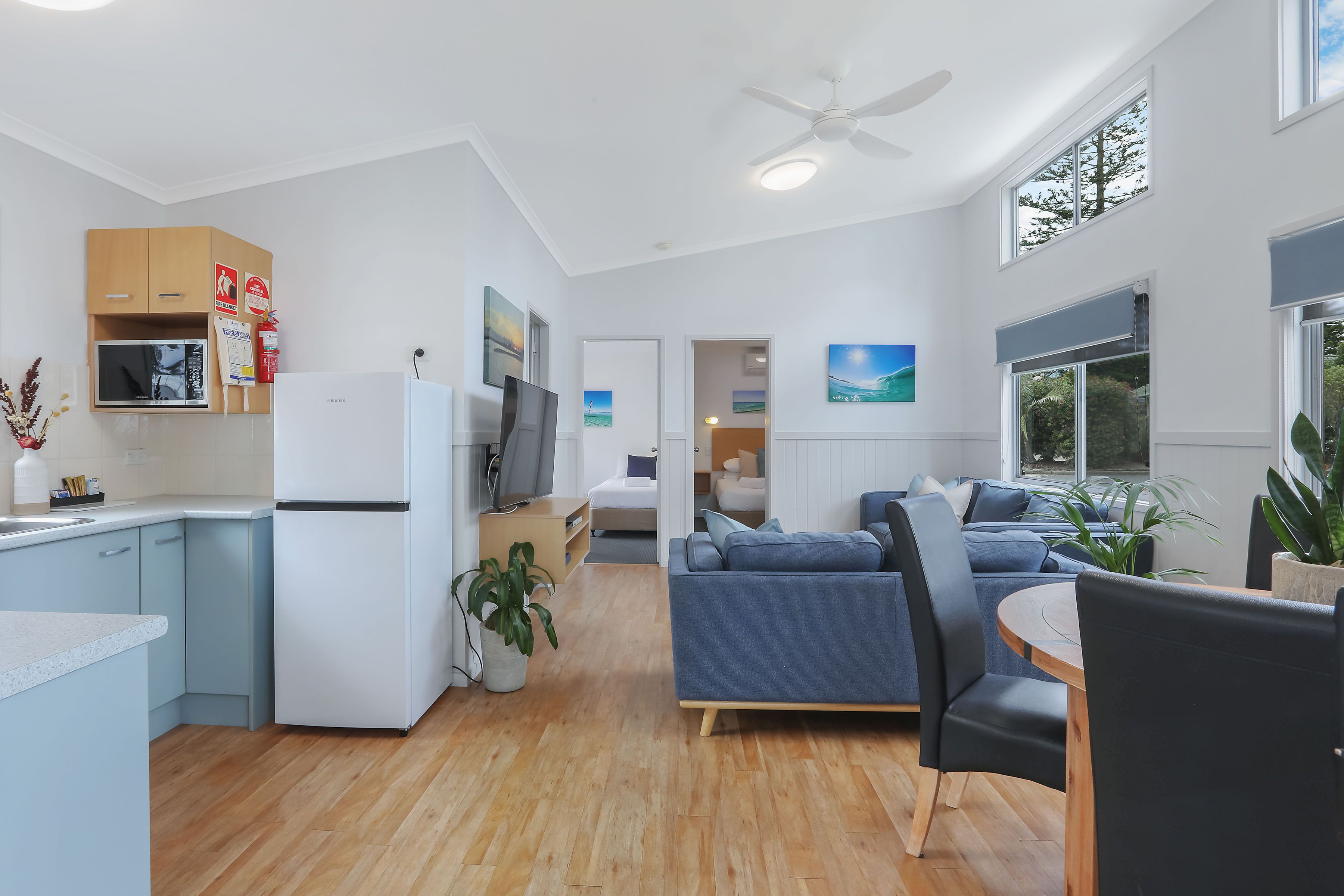 A modern kitchen with a refrigerator, microwave, and cabinets. There is a dining area with a table and chairs, and a living room with a sofa and a TV.
